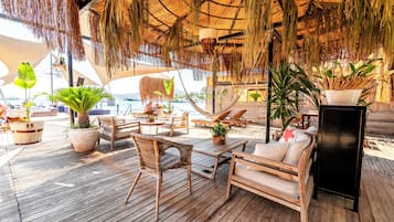 Una terraza de madera con sillones y mesas, rodeada de plantas en macetas y un techo de paja.