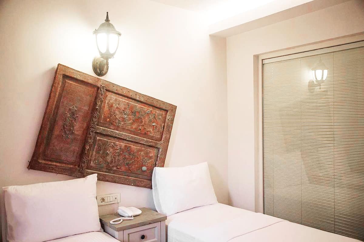 A hotel room with a bed, bedside table, lamp, and window with blinds.
