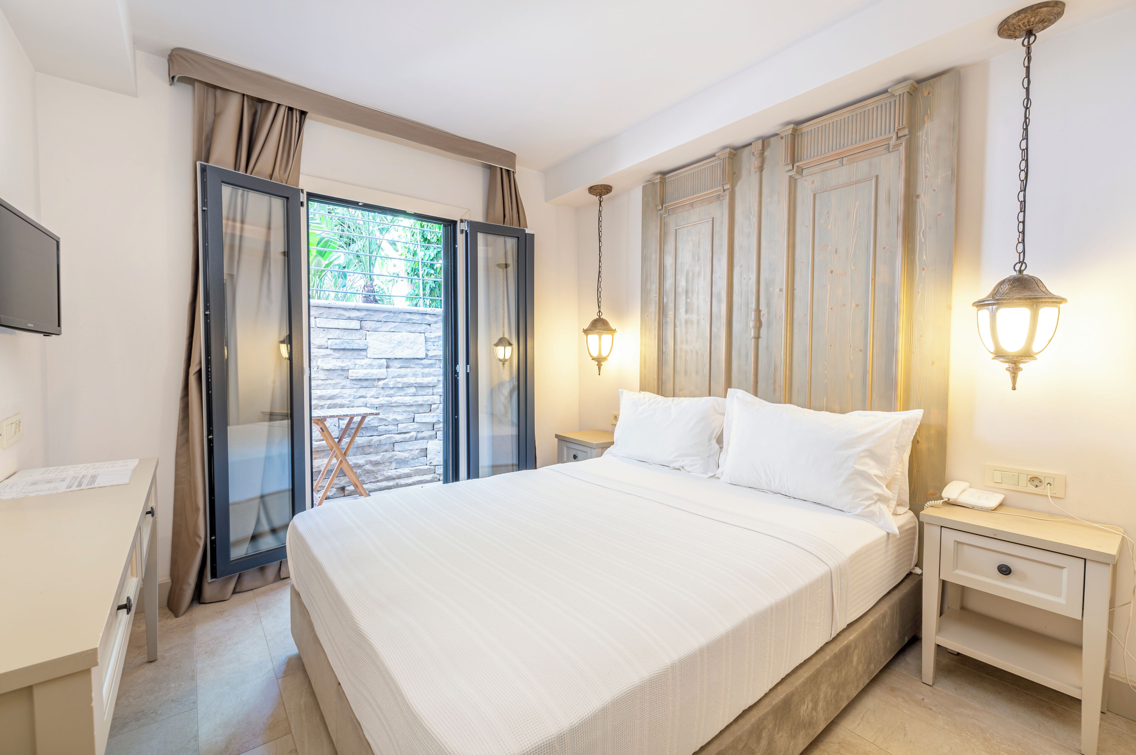 Un dormitorio con cama, mesita de noche, escritorio y puerta corrediza de vidrio que da a un espacio exterior.