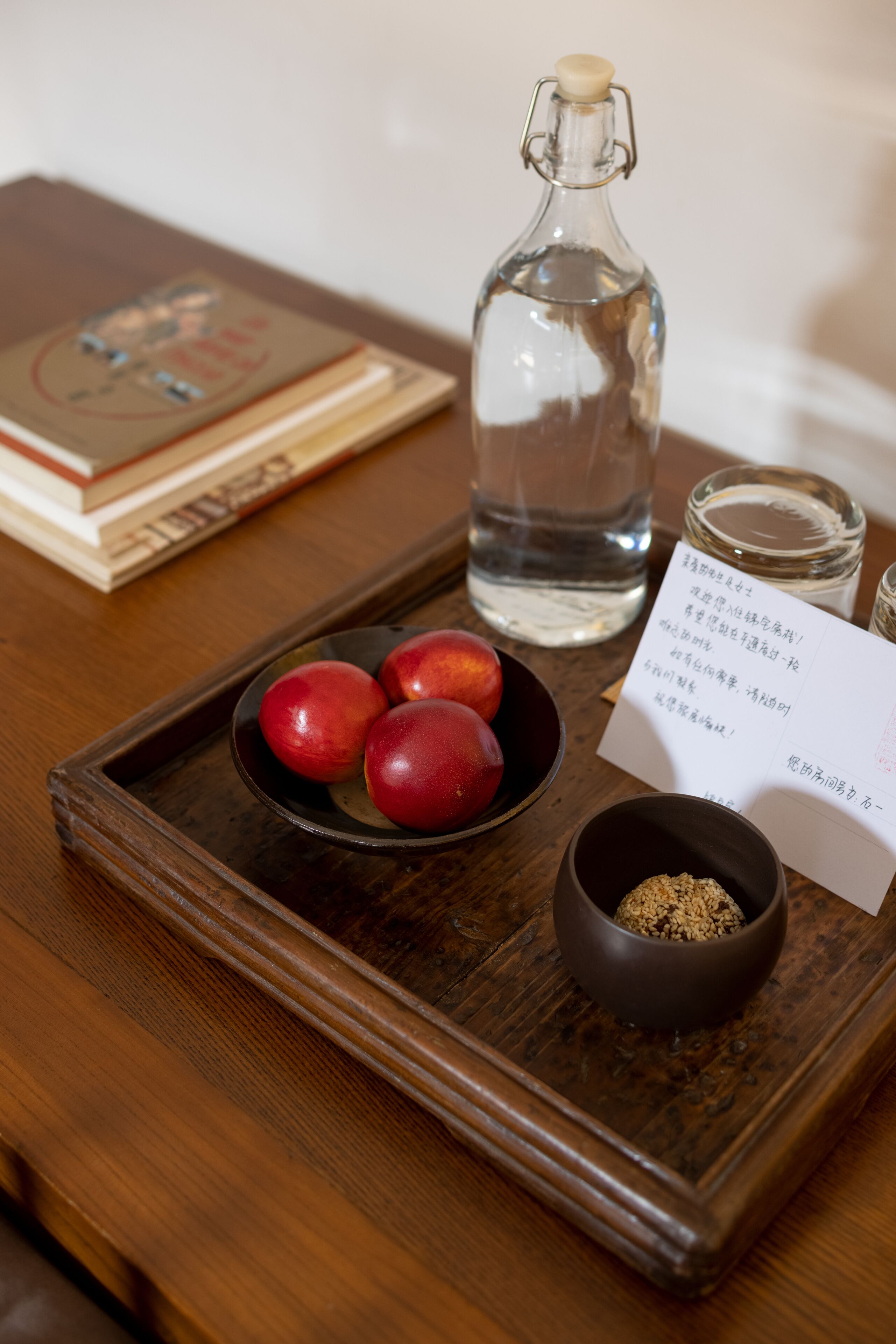 A wooden tray with a bottle, a bowl of red fruits, a bowl of brown substance, and a card.
