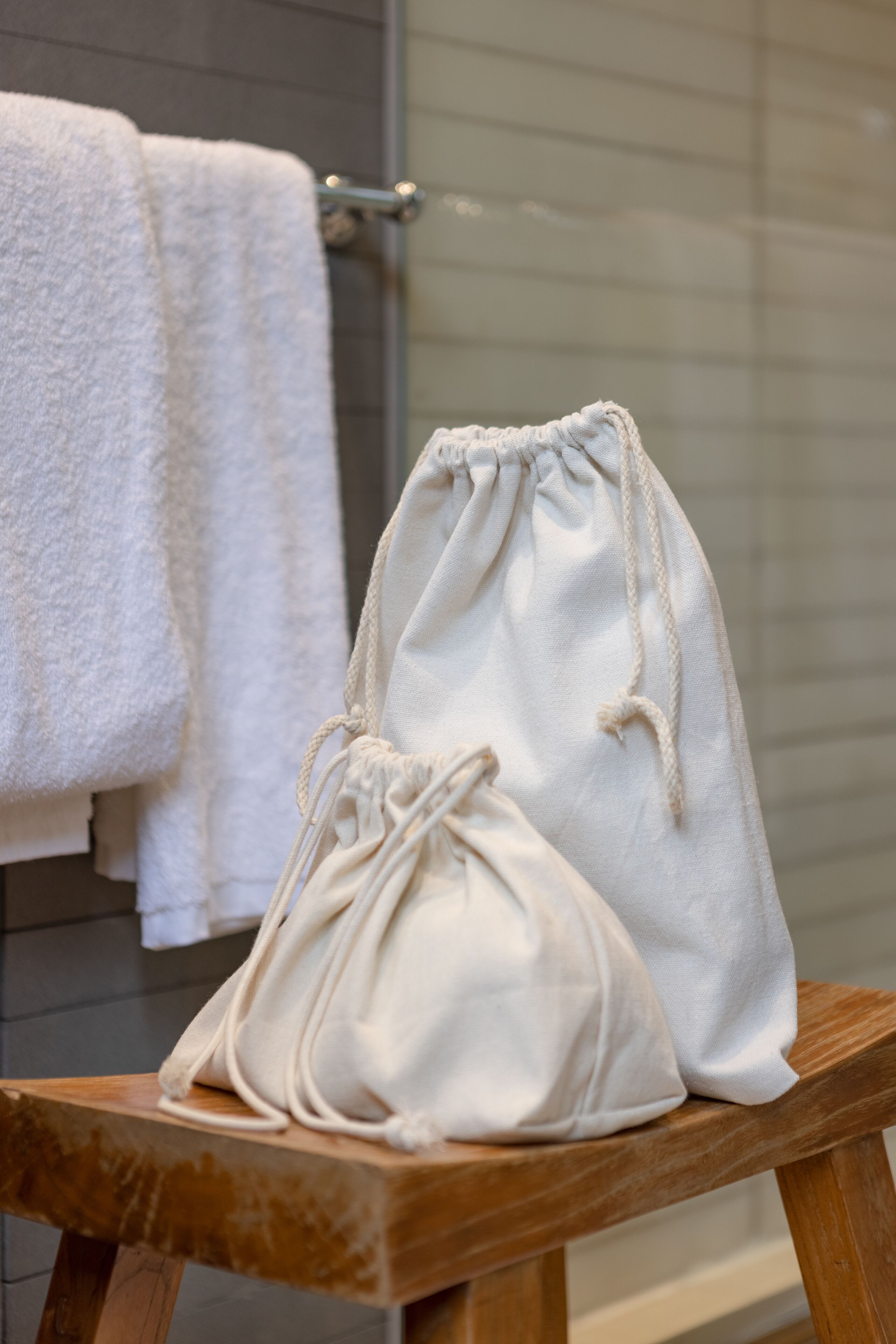 A white drawstring bag on a wooden stool in a bathroom with a white towel hanging on a rack.