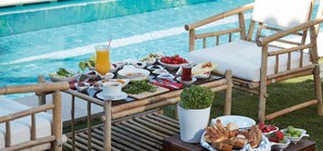 A poolside dining setup with a table set for breakfast, including various dishes, a drink, and a small potted plant.