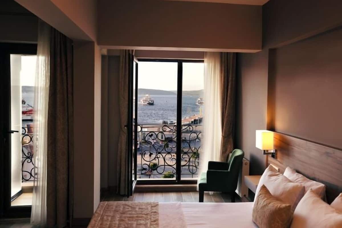 A hotel room with a bed, a chair, and a balcony with a view of the sea and boats.
