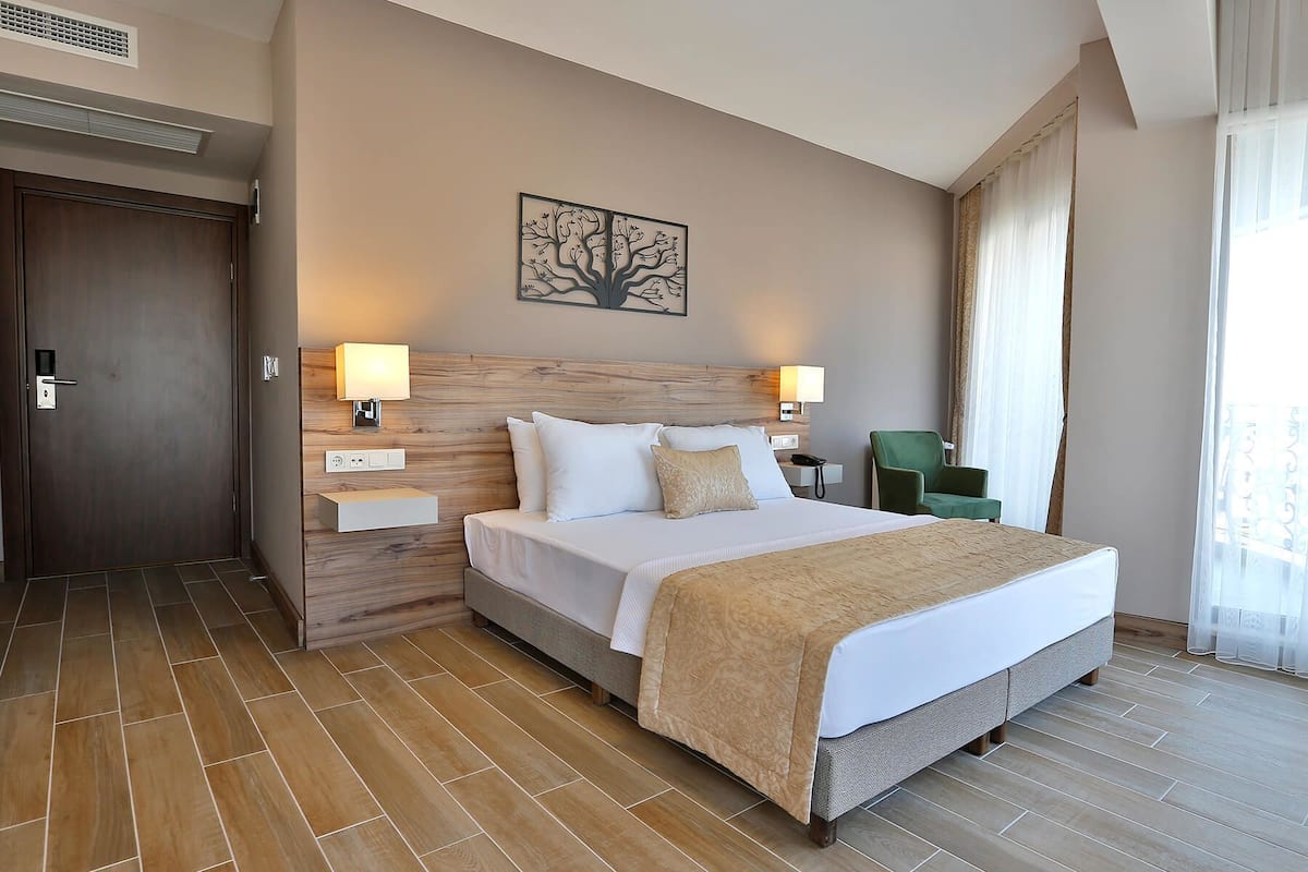 A hotel room with a large bed, two bedside tables, a wall-mounted artwork, a green chair, and a window with curtains.