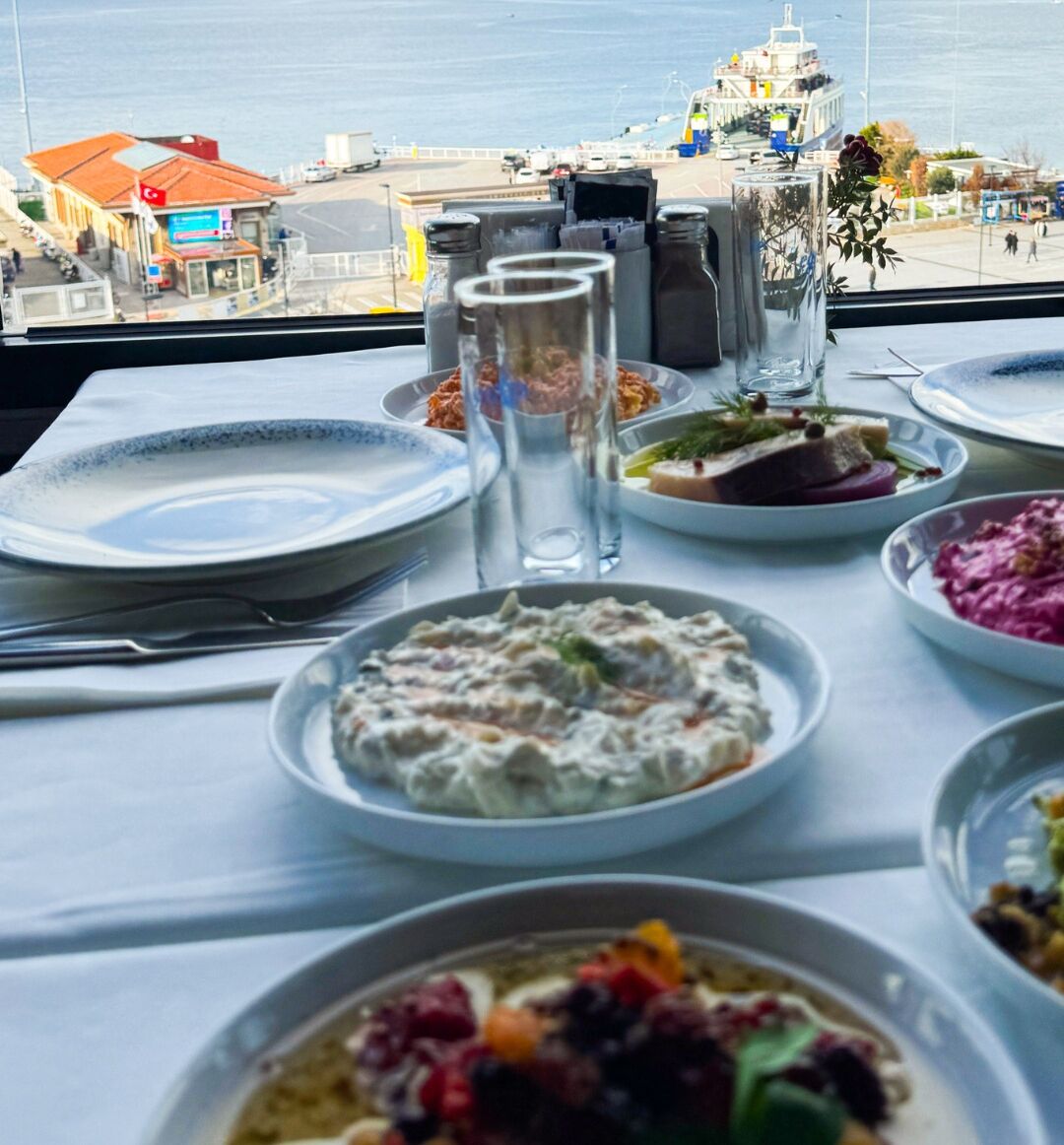 Una mesa dispuesta con varios platos, vista al mar y un barco en el fondo.