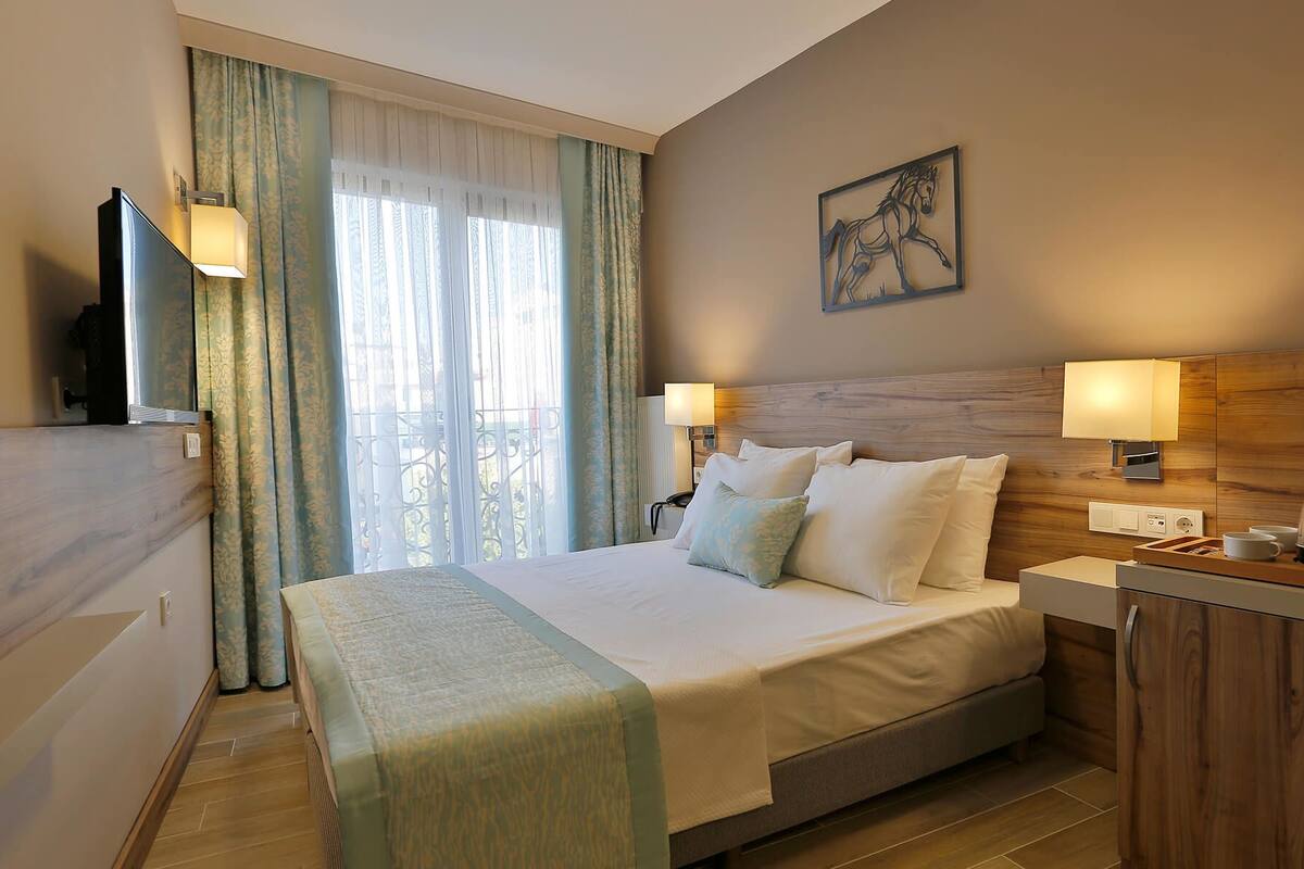 A hotel room with a bed, bedside lamps, a painting, and a view of the outdoors through a window with curtains.