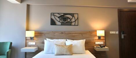 Habitación de hotel con una cama grande, dos mesitas de noche con lámparas, una silla verde, un cabecero de madera y una obra de arte en la pared.