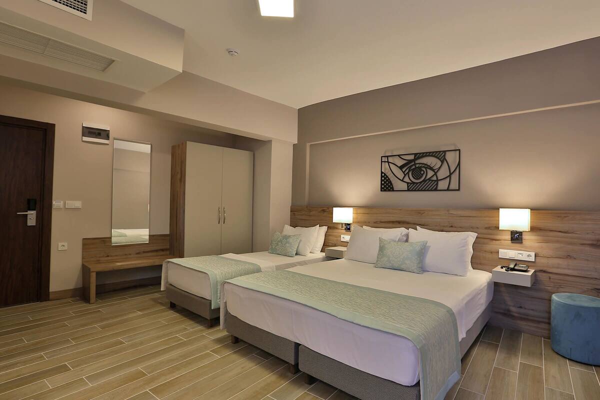A hotel room with two beds, a wooden headboard, a mirror, a wall art piece, and a small blue ottoman.