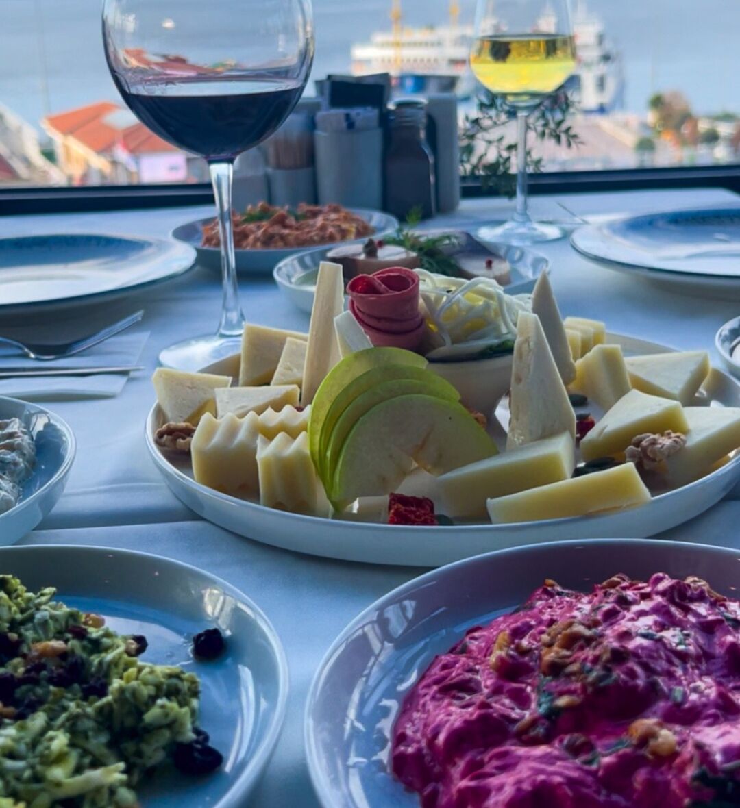 Una mesa servida con varios platos, incluyendo queso, frutas y un bol de ensalada, con una vista de un barco al fondo.