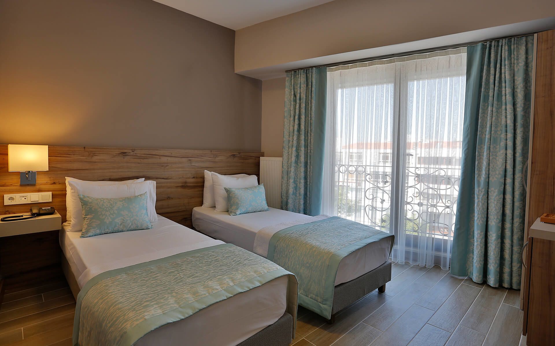 Habitación de hotel con dos camas, cabecero de madera, mesita de noche con lámpara y vista al exterior a través de cortinas traslúcidas.