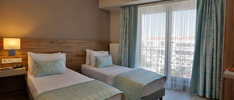 Habitación de hotel con dos camas, cabecero de madera, mesita de noche con lámpara y vista al exterior a través de cortinas traslúcidas.