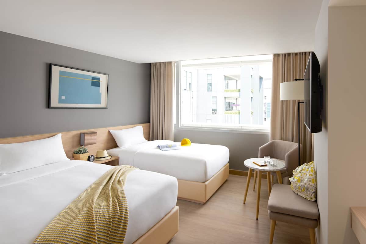 A hotel room with two beds, a small table, a chair, a TV, and a window with a city view.