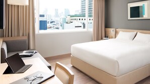 A hotel room with a bed, a desk with a laptop, a chair, a TV, and a window with a city view. - KritThai Residence (Bangkok)