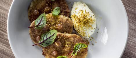 Un plato blanco con alimentos, incluyendo un huevo pochado, un trozo de carne y un pan plano, adornado con hierbas y especias.