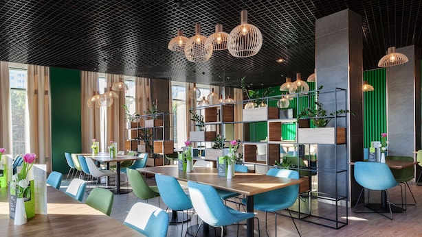 A modern dining area with wooden tables and blue chairs, surrounded by green walls and potted plants. - Hampton by Hilton Lublin (Lublin)