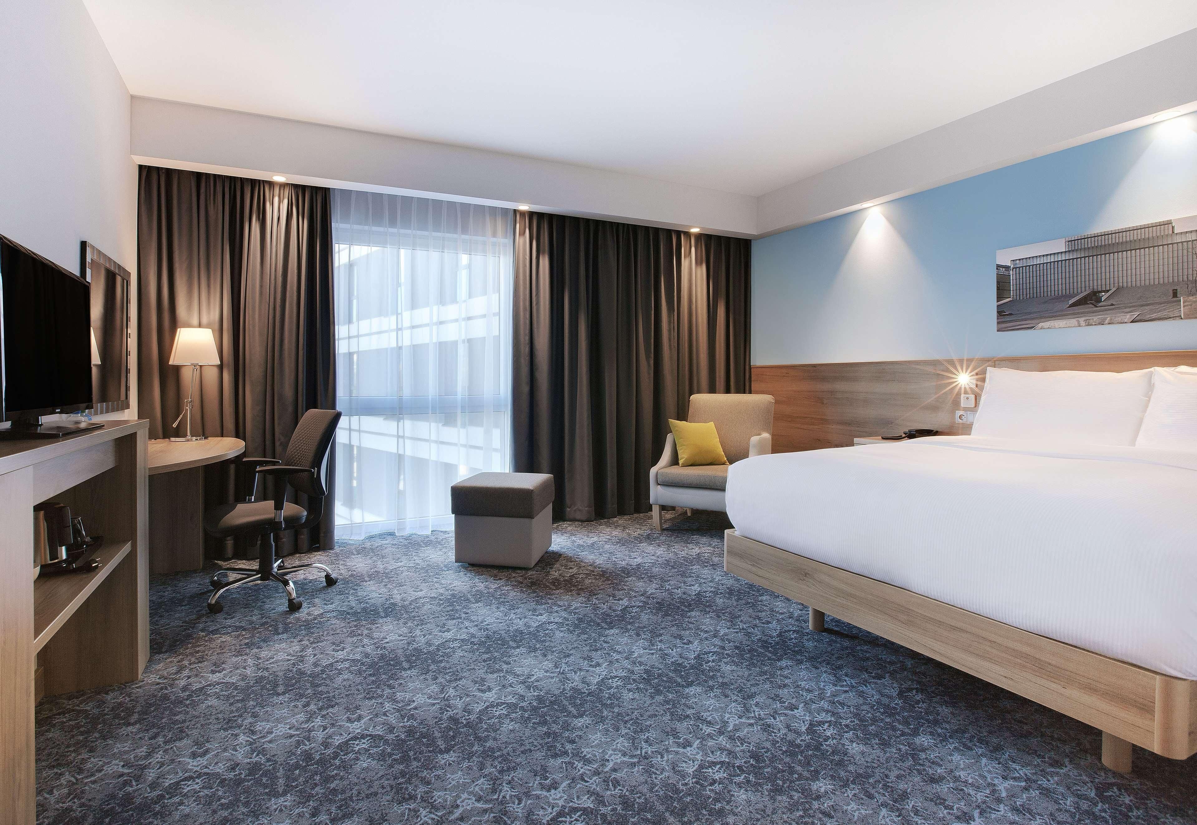 A hotel room with a bed, desk, chair, ottoman, and a large window with curtains.