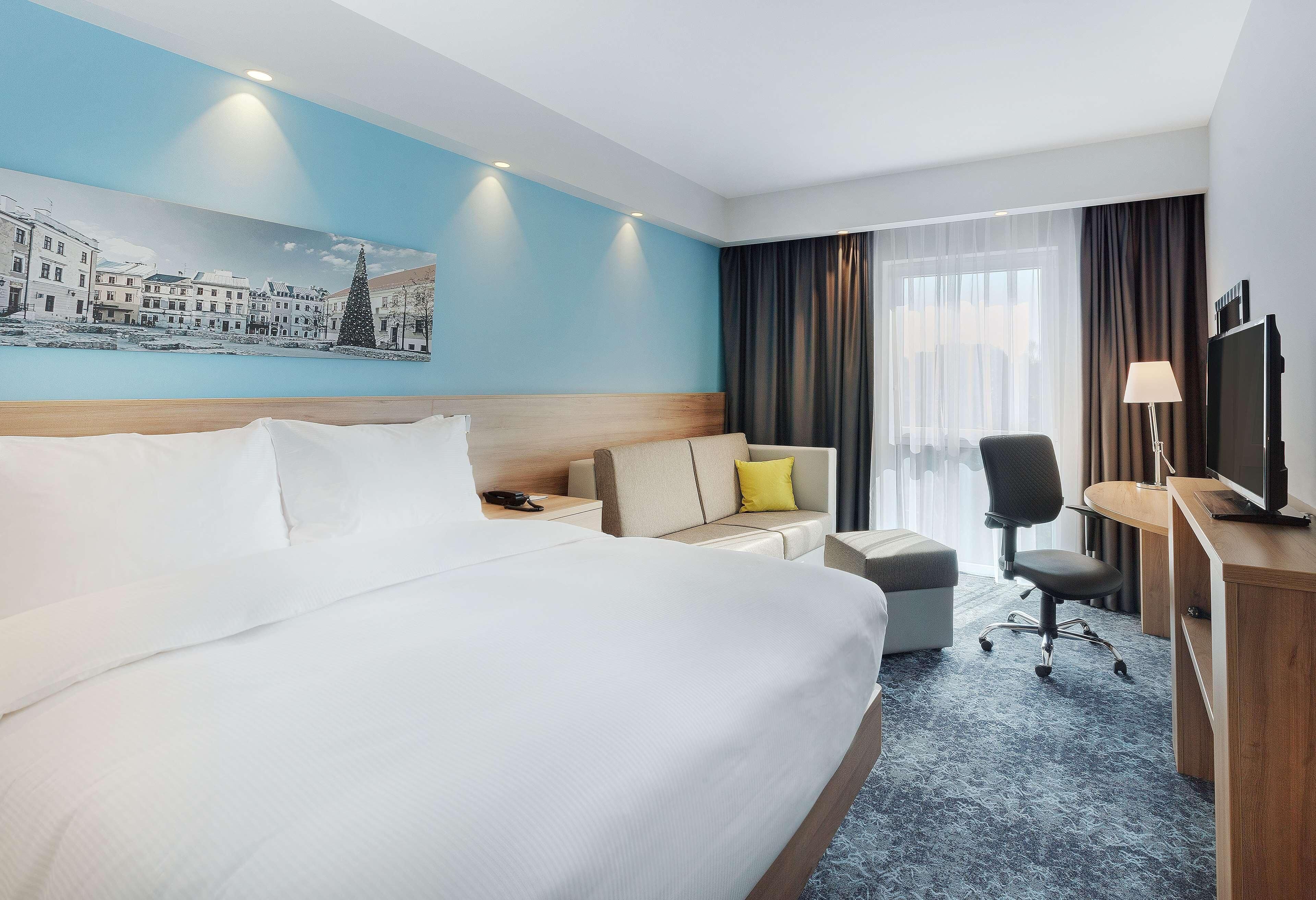 A hotel room with a large bed, a desk, a chair, a sofa, and a television.