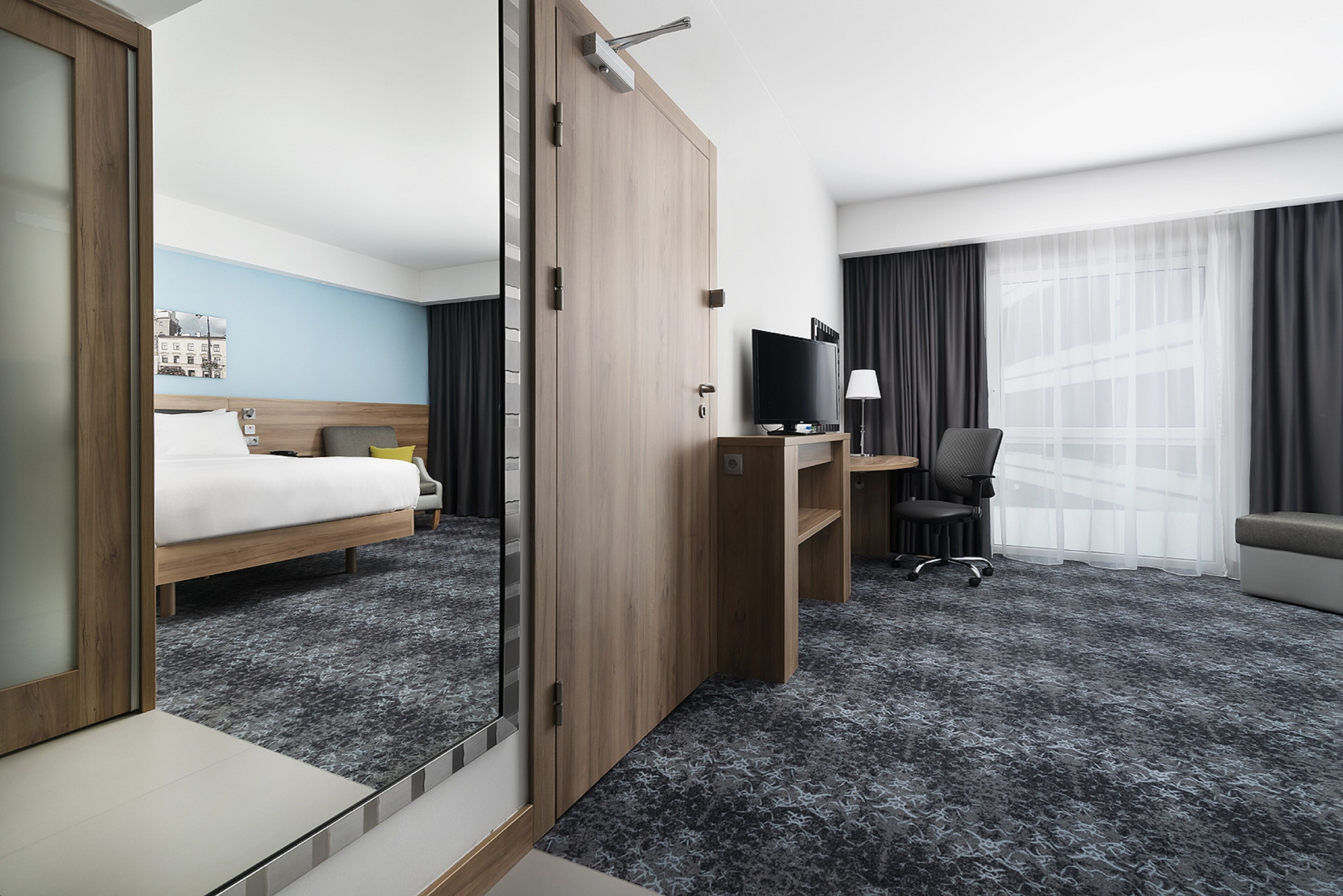 A hotel room with a bed, a desk with a TV, a chair, and a large window with curtains.