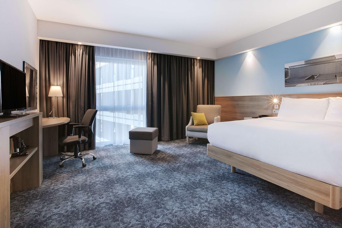 Una habitación de hotel con cama, escritorio, silla, reposapiés y un ventanal con cortinas.