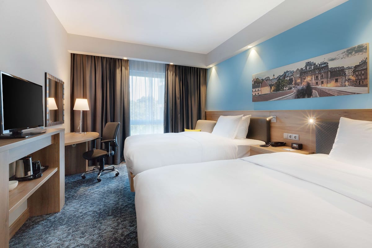 Une chambre d’hôtel avec deux lits, un bureau avec une télévision, une chaise et une grande fenêtre avec des rideaux.