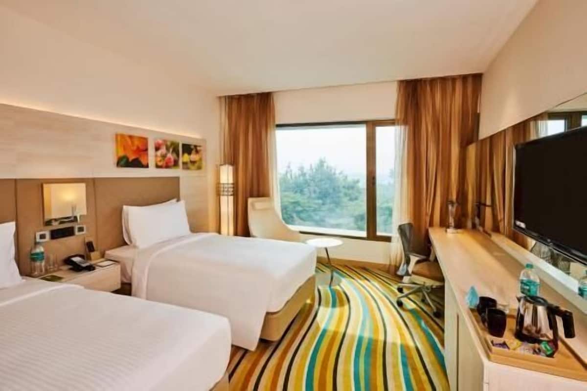 A hotel room with two beds, a desk, a TV, and a window with a view of trees.