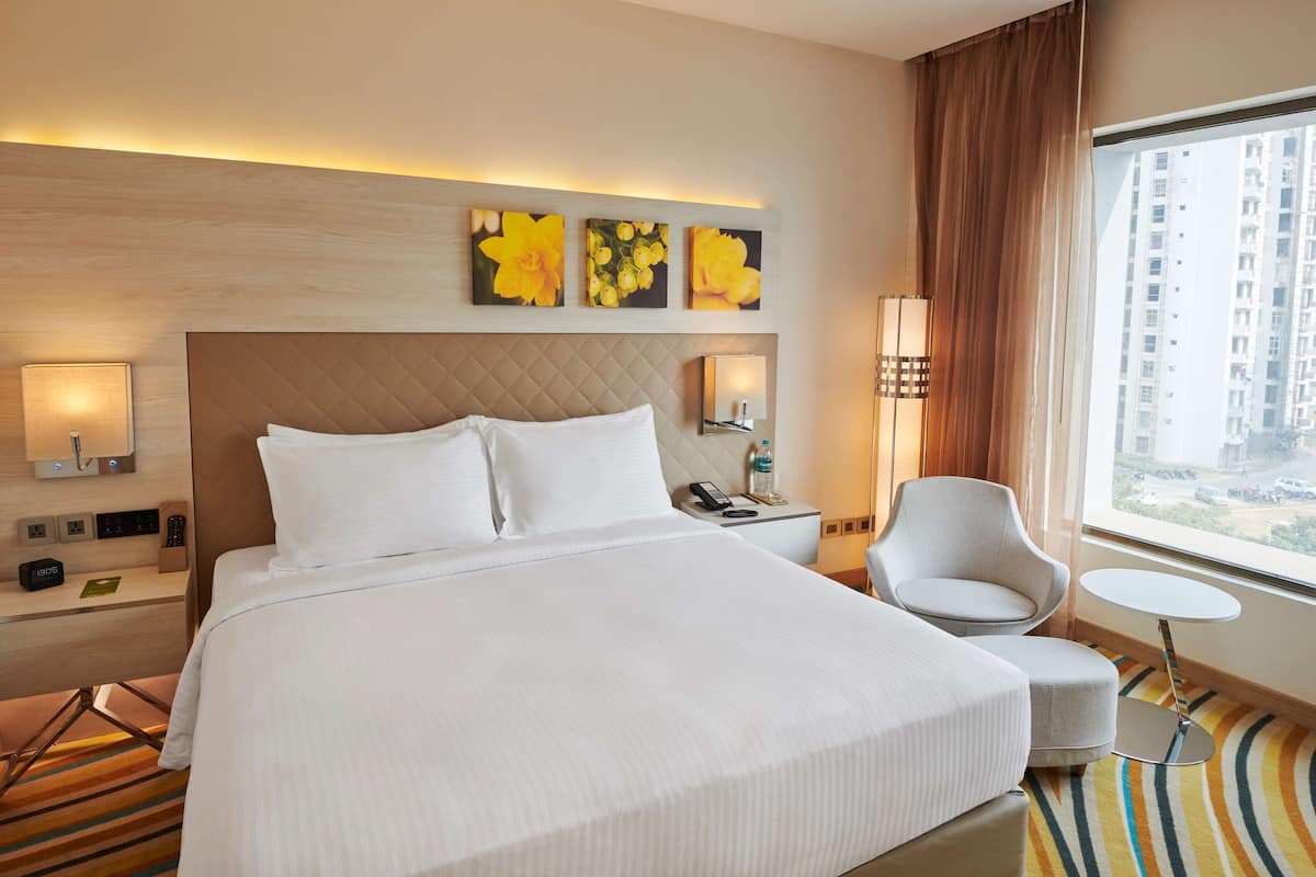 A hotel room with a large bed, a bedside table with a lamp, a chair, a small table, and a view of buildings through the window.