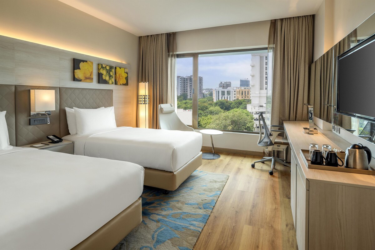 Ein Hotelzimmer mit zwei Betten, einem Schreibtisch mit Stuhl, einem großen Fenster mit Blick auf die Stadt und einem Flachbildfernseher.