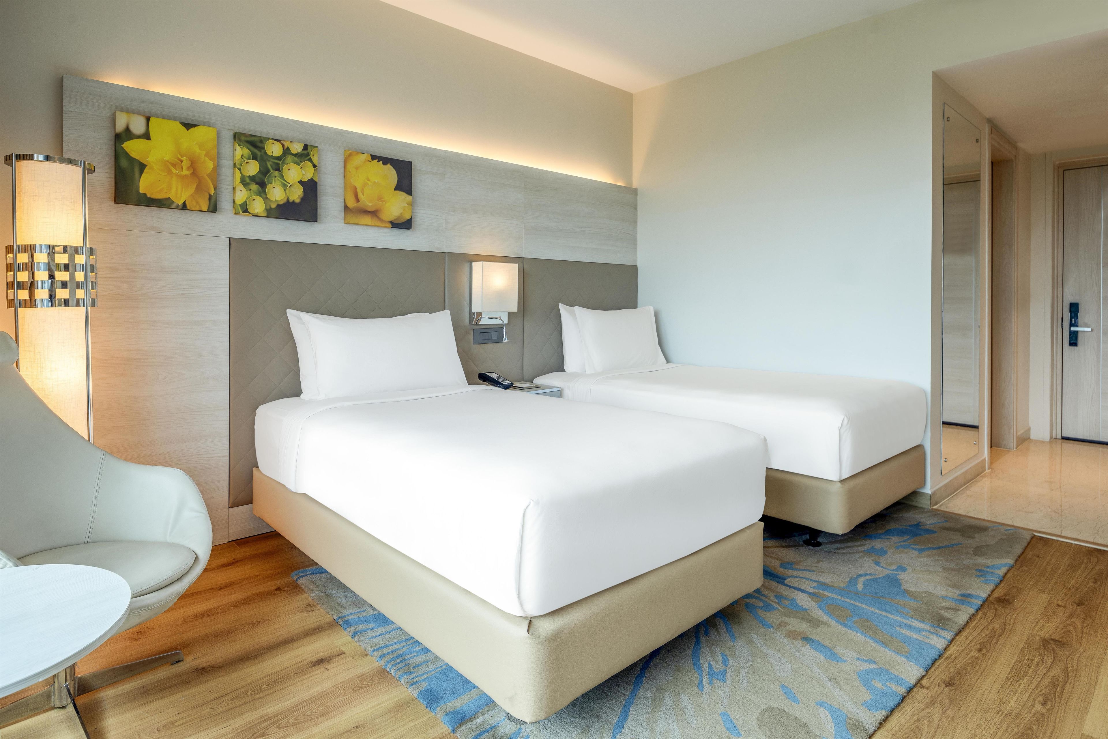 A hotel room with two beds, a desk, a chair, and a wall-mounted lamp.