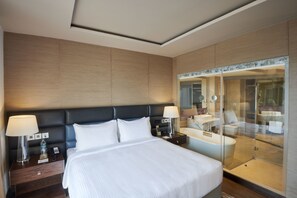 Ein modernes Hotelzimmer mit einem großen Bett, Nachttischen, einem Beistelltisch, einem Glaskastenbad und einem Spiegel.