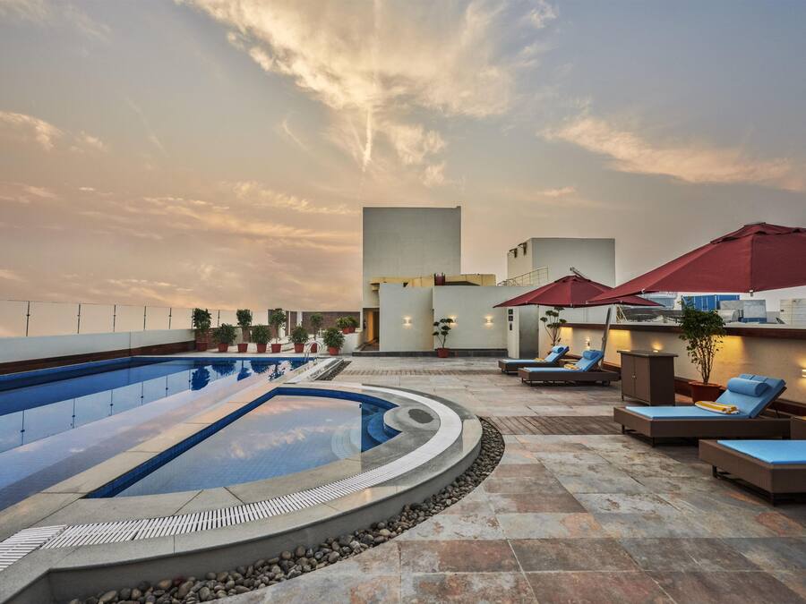 Un área de piscina en la azotea con sillones de descanso y sombrillas, rodeada de plantas en macetas y una estructura de edificio moderna.