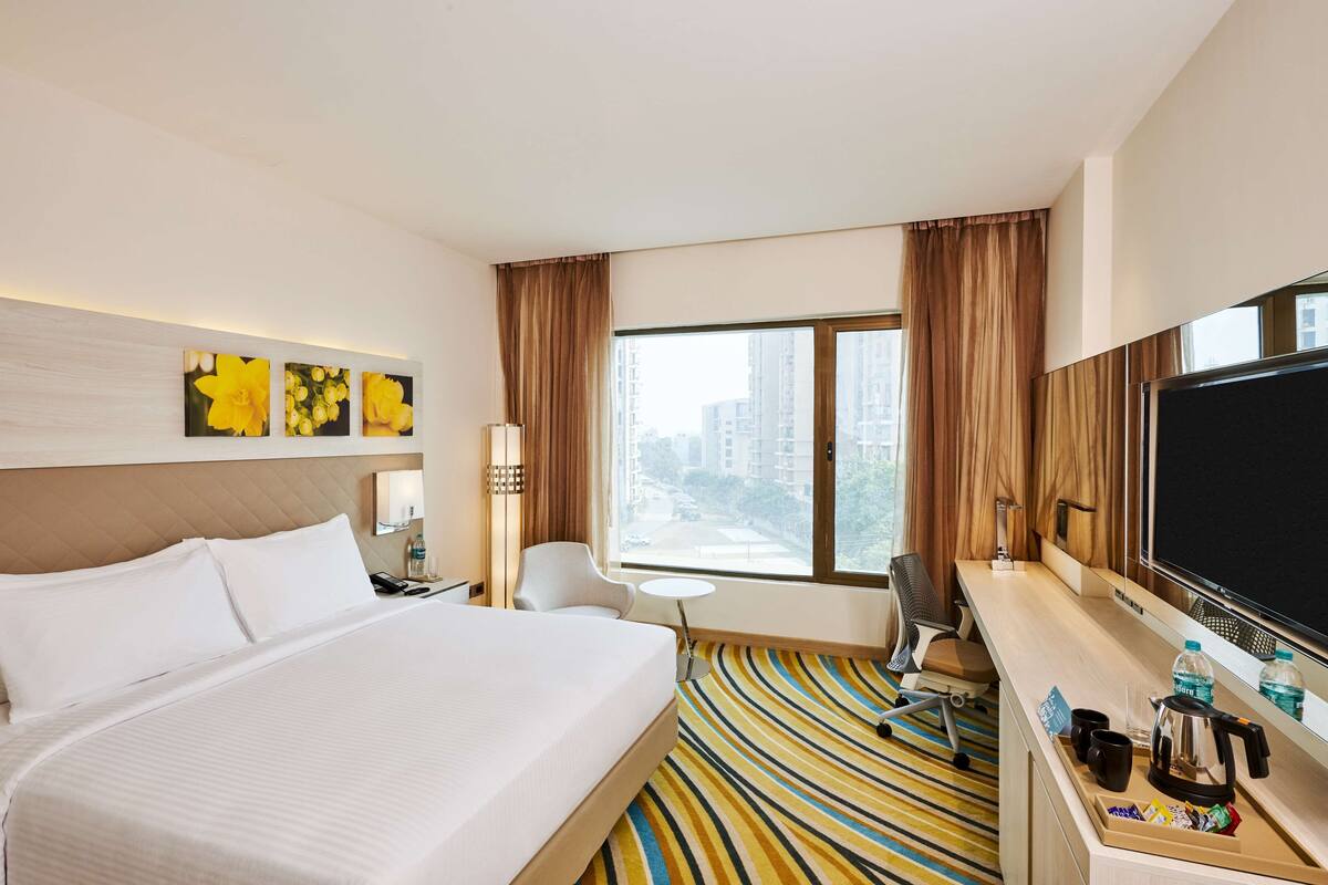A hotel room with a large bed, a desk with a coffee maker, a TV, and a view of buildings through the window.