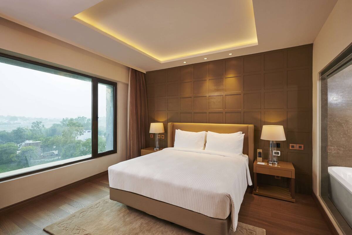 A modern hotel room with a large bed, two bedside tables with lamps, a view of the city, and a large window.