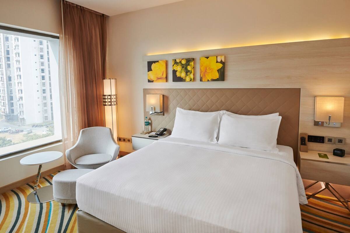 A hotel room with a large bed, a chair, a small table, a bedside lamp, a phone, and artwork on the wall.