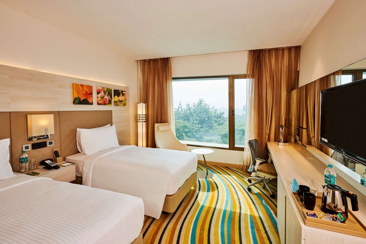 A hotel room with two beds, a desk with a TV, a desk lamp, a chair, a coffee maker, and a window with a view of trees.