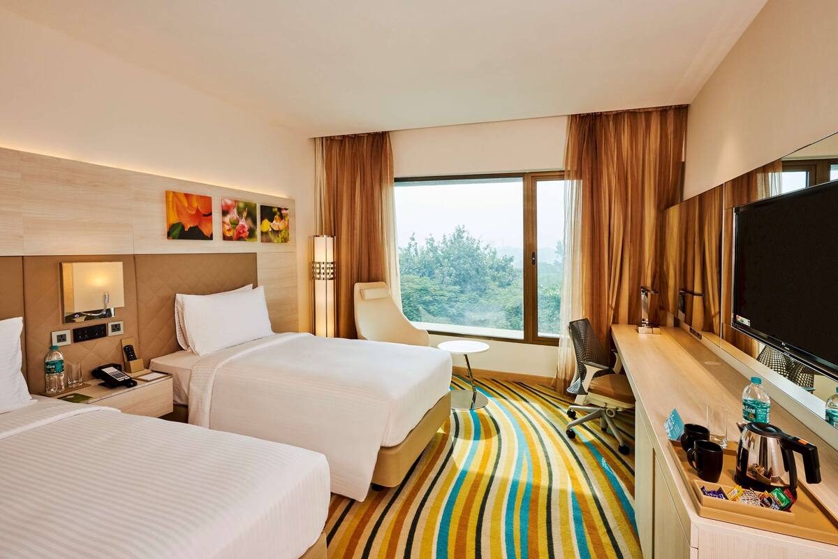 A hotel room with two beds, a desk with a flat-screen TV, a window with curtains, and a striped carpet.