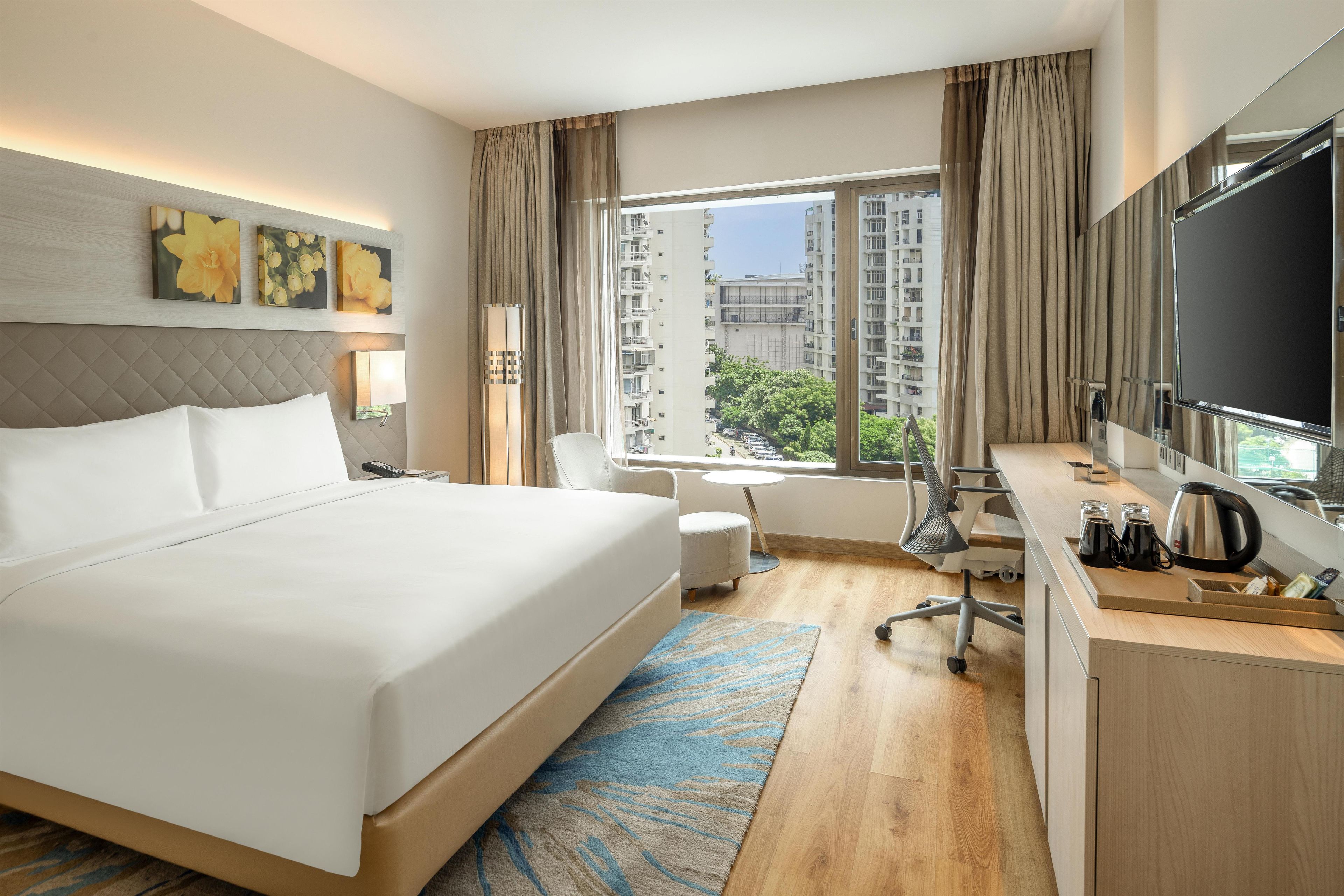 A hotel room with a large bed, a desk with a chair, a TV, and a window with a view of buildings.
