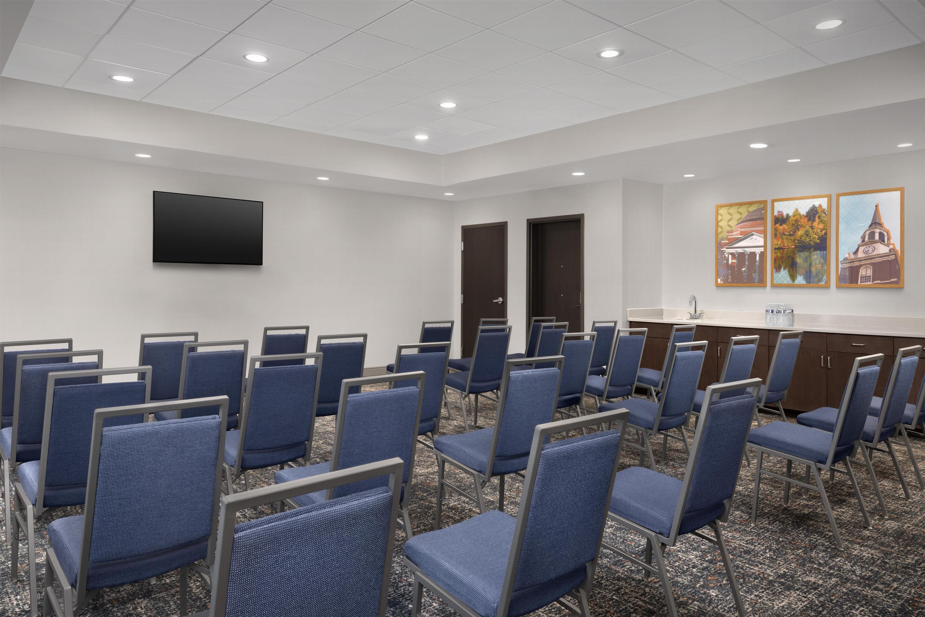Un salón de conferencias con filas de sillas azules, un televisor de pantalla plana y cuadros en la pared.