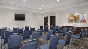 Un salón de conferencias con filas de sillas azules, un televisor de pantalla plana y cuadros en la pared.