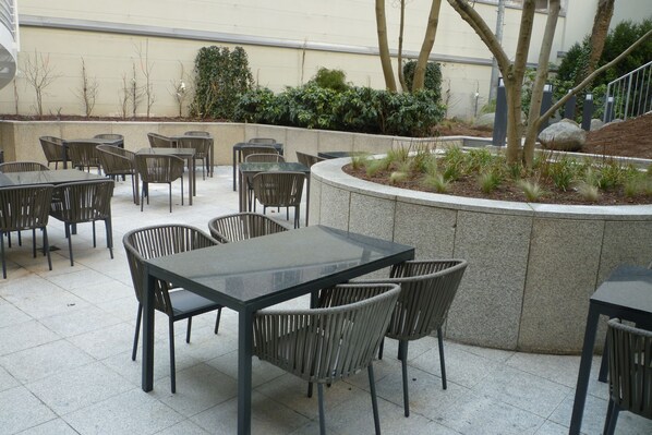 Un patio al aire libre con varias mesas y sillas, rodeado por un muro de piedra bajo y vegetación.