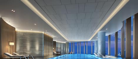 Piscina interior moderna con vistas despejadas al cielo a través de amplios ventanales, rodeada de zonas de descanso con toallas.