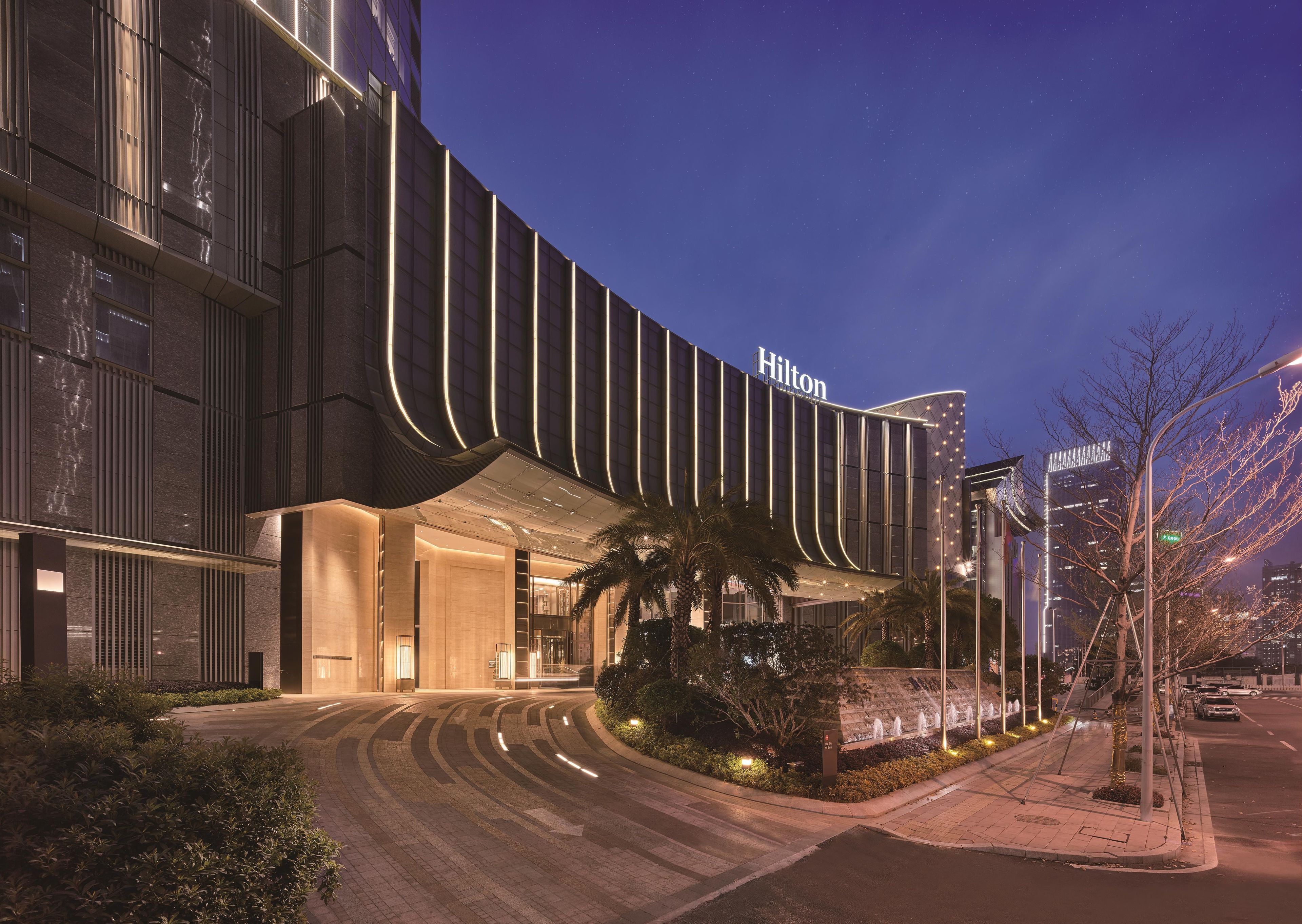 A modern hotel with a curved facade, illuminated signage, and a landscaped entrance area.