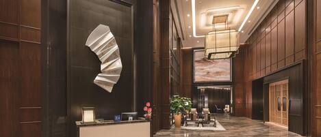 A modern hotel lobby with a reception desk, seating area, and a large decorative wall piece.