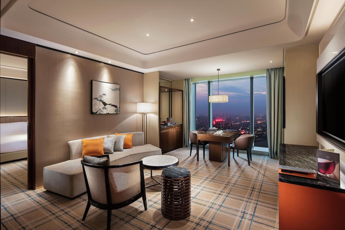 A modern hotel room with a sofa, dining table, chairs, a small round table, a lamp, a TV, and a view of the city at night.