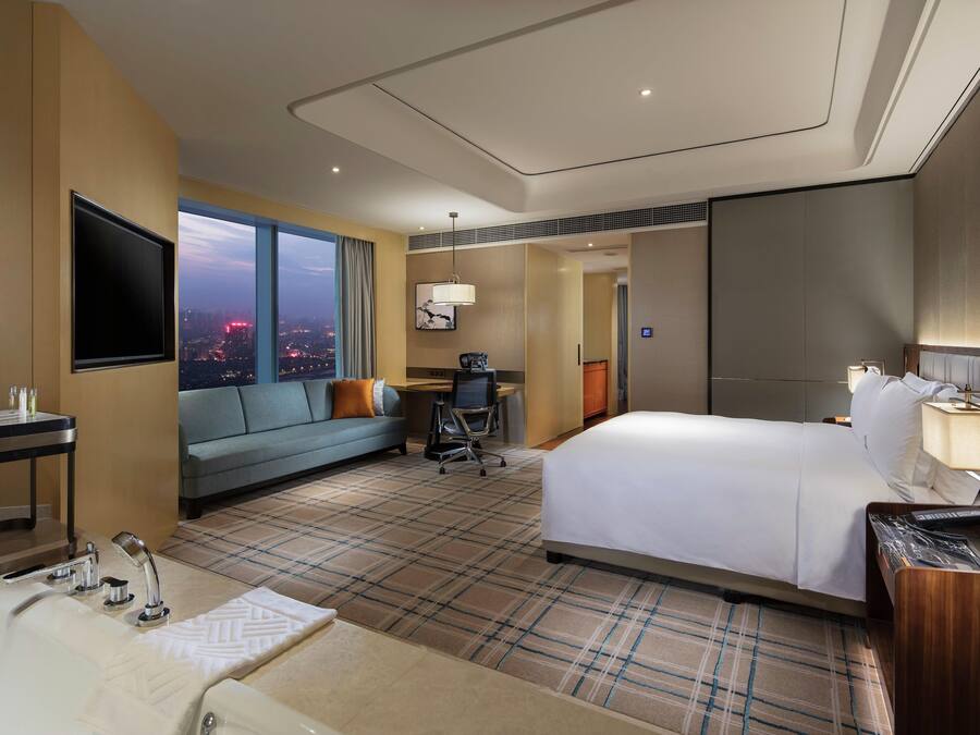 Een hotelkamer met een groot bed, een slaapbank, een bureau, een televisie en uitzicht op de stad in de schemering.