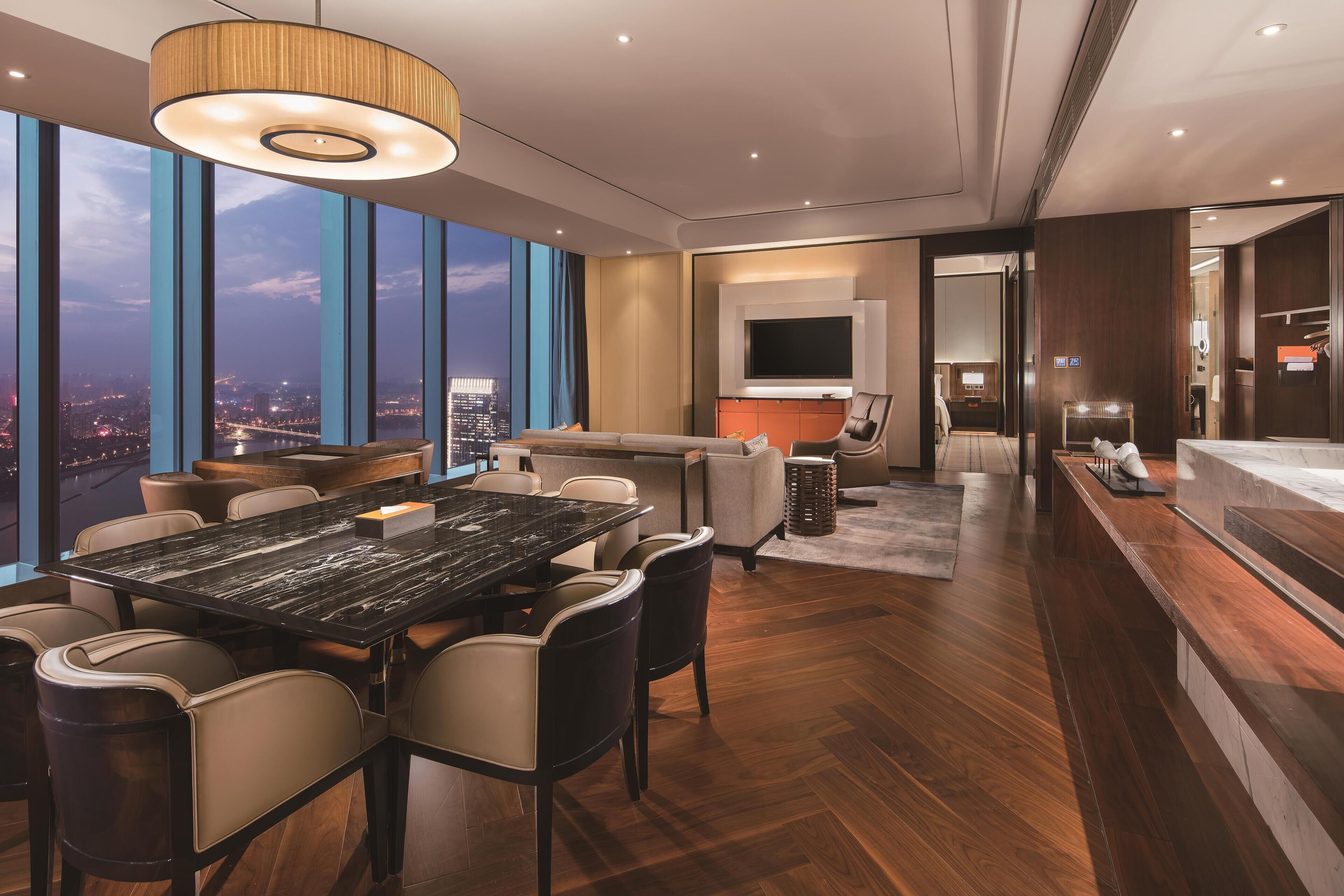 Une chambre d’hôtel moderne dotée d’une grande table à manger, de sièges confortables, d’un téléviseur et offrant une vue sur le paysage urbain.