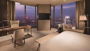 Habitación de hotel con una cama grande, un escritorio con una silla, un televisor montado en la pared y vistas a la ciudad al anochecer.
