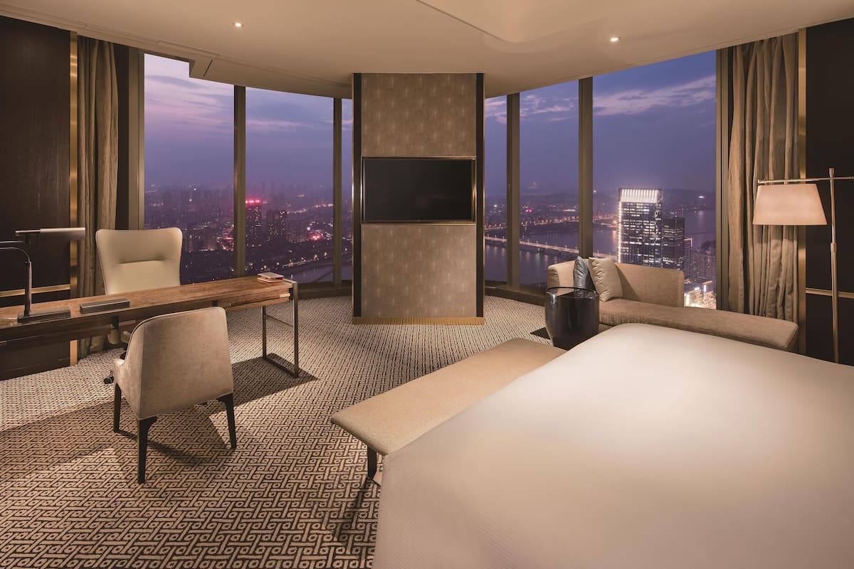 A hotel room with a large bed, a desk with a chair, a TV mounted on the wall, and a view of the city at dusk.