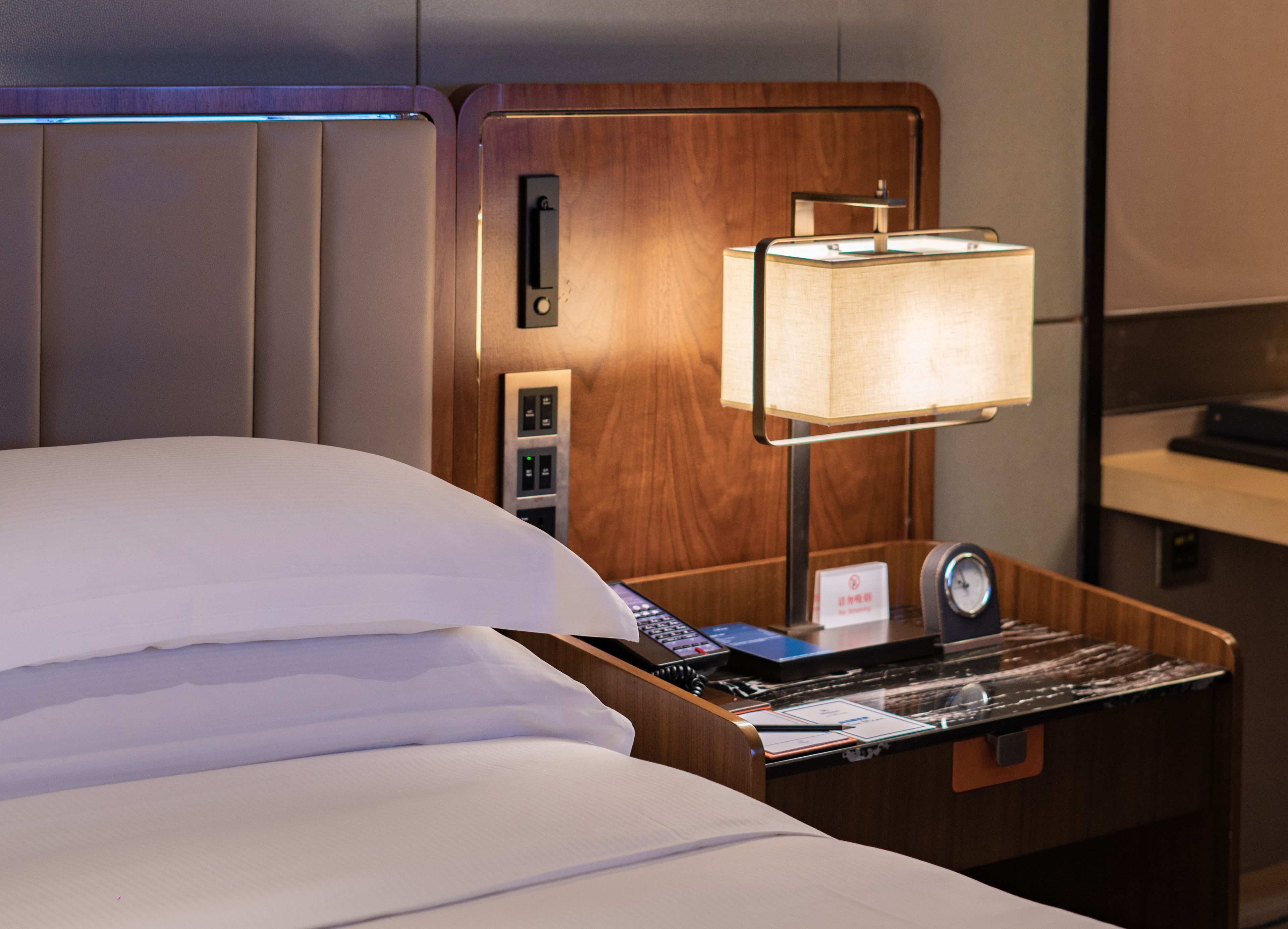 Quarto de hotel com cama, mesa de cabeceira com luminária, relógio e painel de controle.