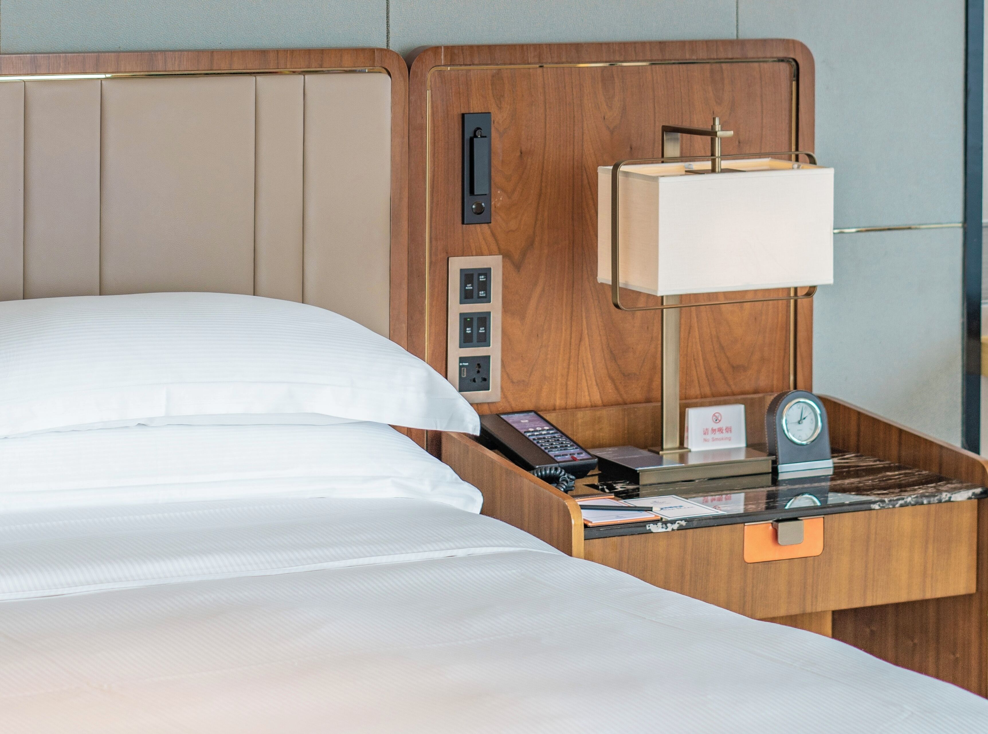 Habitación de hotel con cama, controles junto a la cama, un reloj y una mesita de noche con lámpara y un cartel.