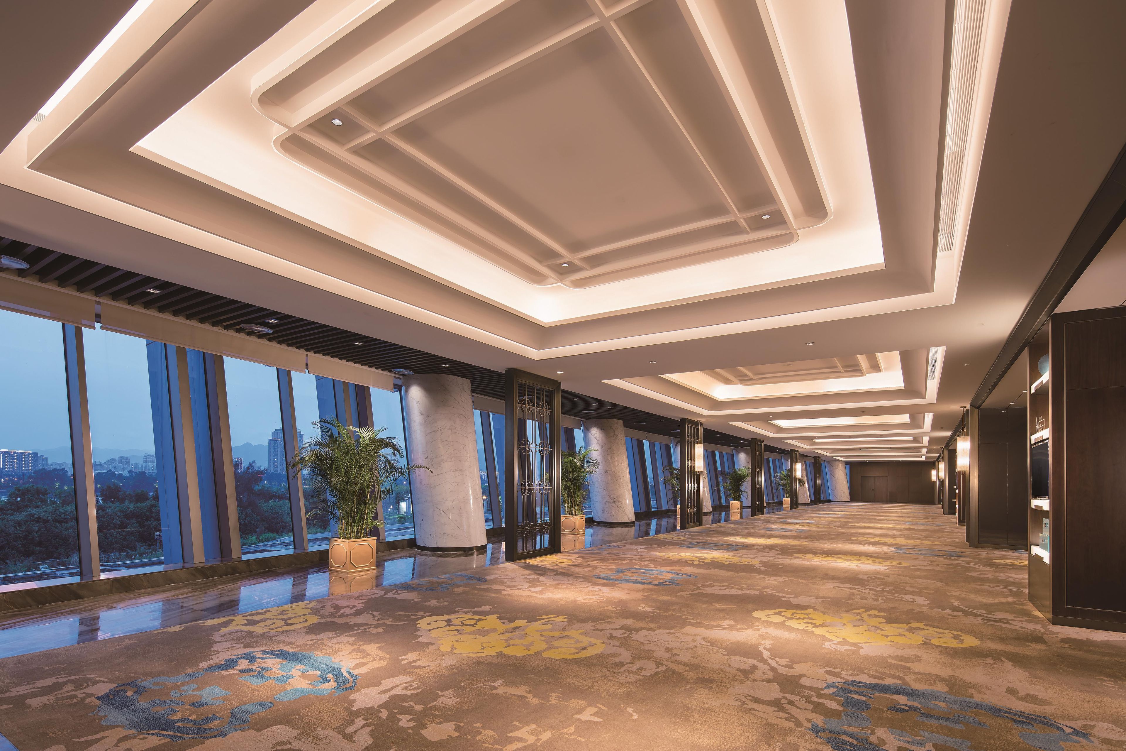Un vaste couloir d’hôtel, doté de grandes fenêtres, d’un tapis à motifs et de luminaires modernes.